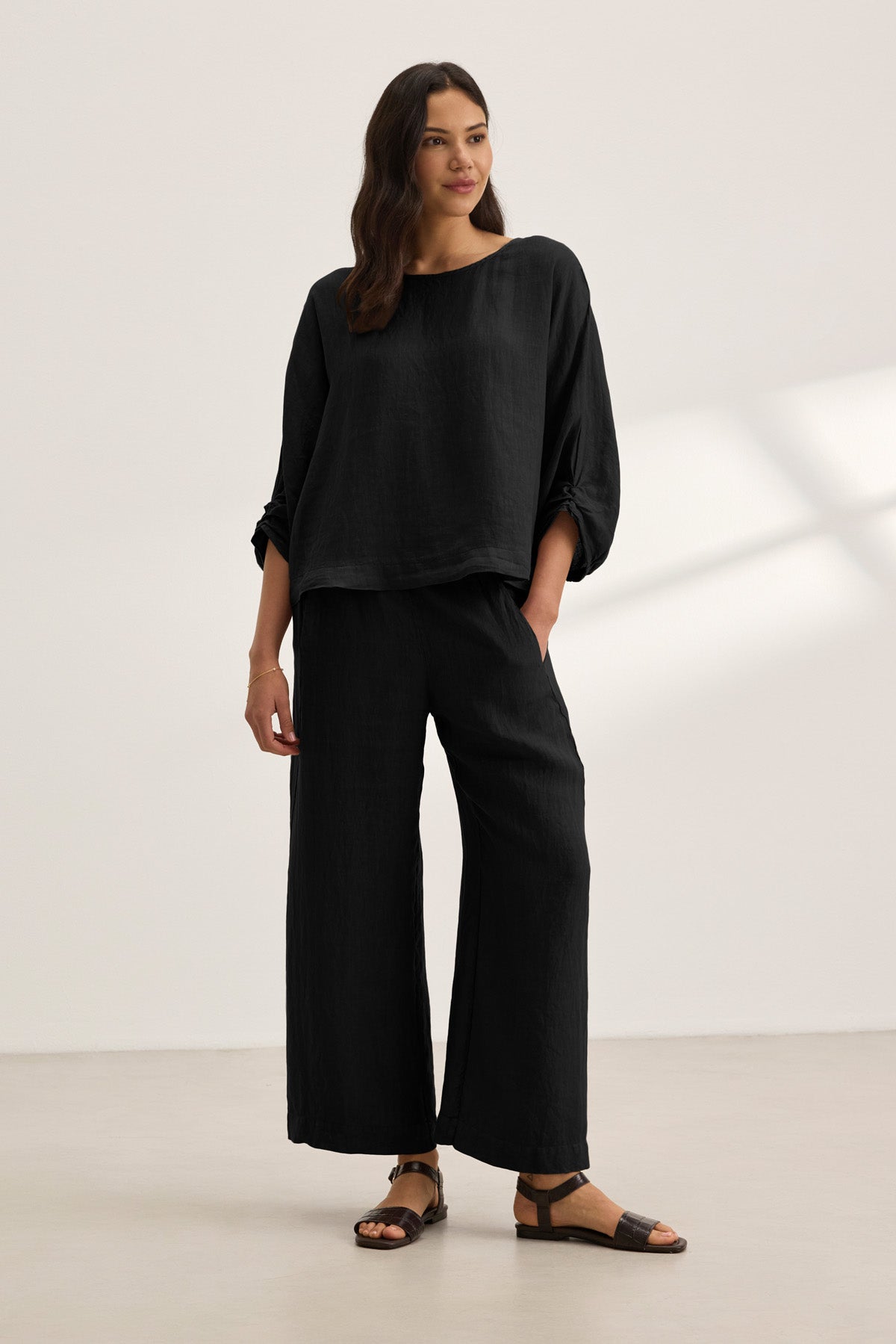 A woman stands indoors against a plain light background wearing the Velvet by Graham & Spencer CASS LINEN TOP, a loose-fitting black woven linen top with a cocoon silhouette, paired with wide-leg black pants and black sandals.-41160212250817