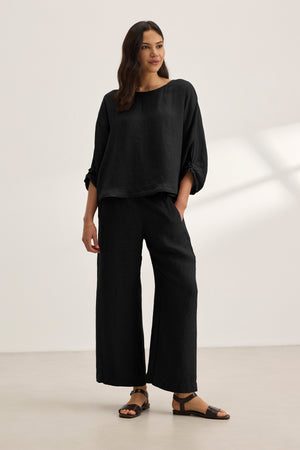 A woman stands indoors against a plain light background wearing the Velvet by Graham & Spencer CASS LINEN TOP, a loose-fitting black woven linen top with a cocoon silhouette, paired with wide-leg black pants and black sandals.