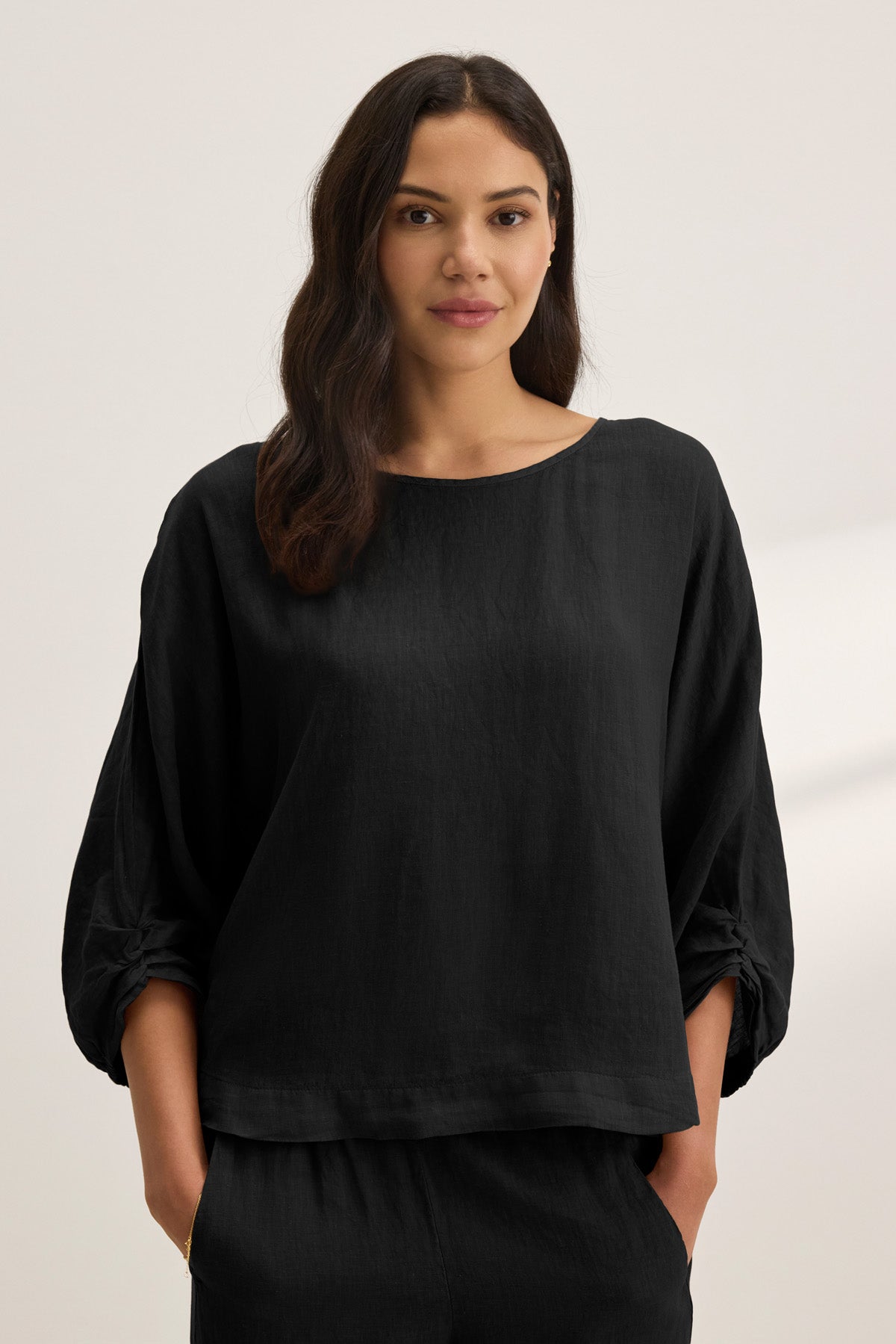 A woman with long, dark hair stands against a plain light background, wearing the Velvet by Graham & Spencer CASS LINEN TOP—an easy-fitting black woven linen piece—with her hands in her pockets.-41160212218049