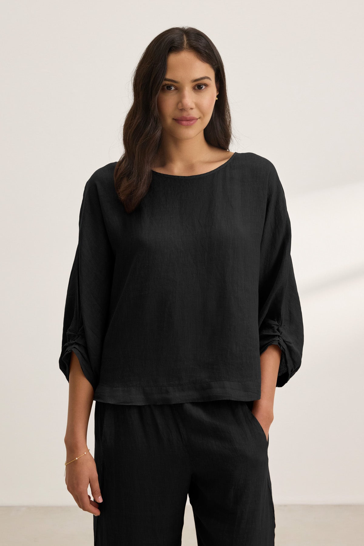 A woman stands against a plain background wearing the CASS LINEN TOP by Velvet by Graham & Spencer—a loose-fitting black linen top with a cocoon silhouette—paired with matching black pants, one hand in her pocket.