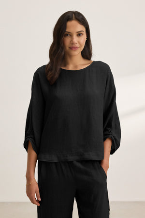 A woman stands against a plain background wearing the CASS LINEN TOP by Velvet by Graham & Spencer—a loose-fitting black linen top with a cocoon silhouette—paired with matching black pants, one hand in her pocket.