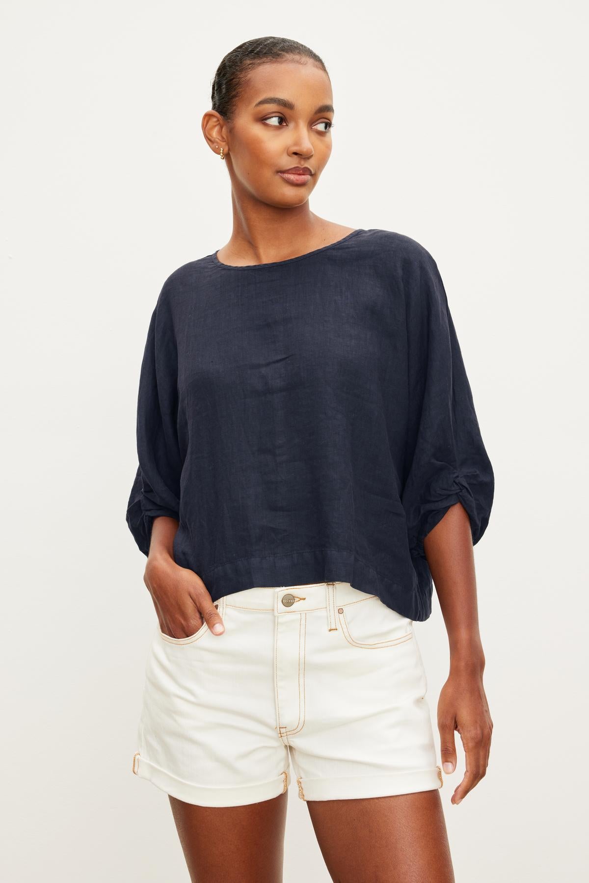   Someone wearing the CASS LINEN TOP, a black long-sleeve piece by Velvet by Graham & Spencer, paired with white shorts stands against a plain background. 