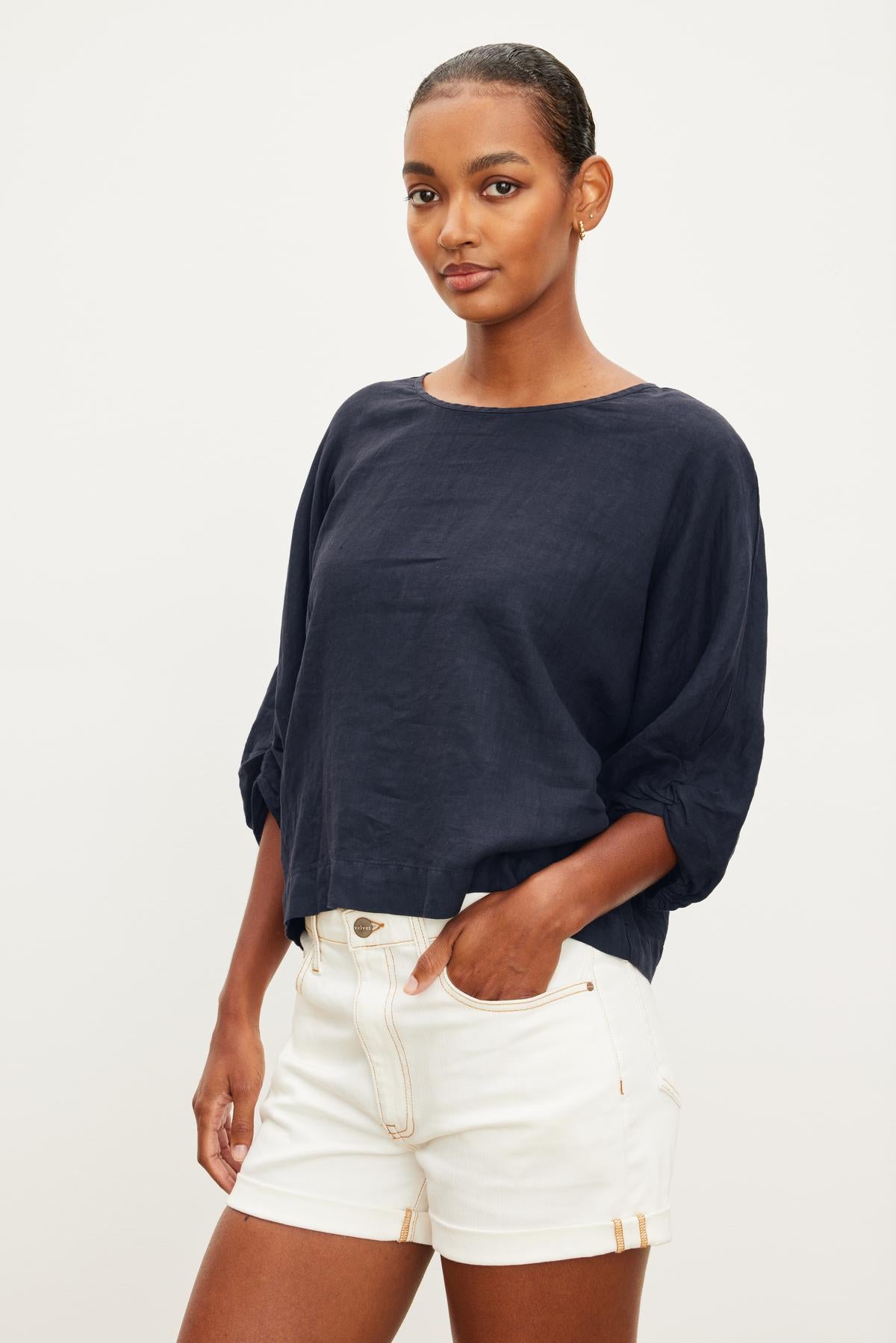   Someone wears the Velvet by Graham & Spencer CASS LINEN TOP in dark blue, featuring a twist knot detail. They pair it with white shorts and stand against a plain backdrop, one hand resting in their pocket. 