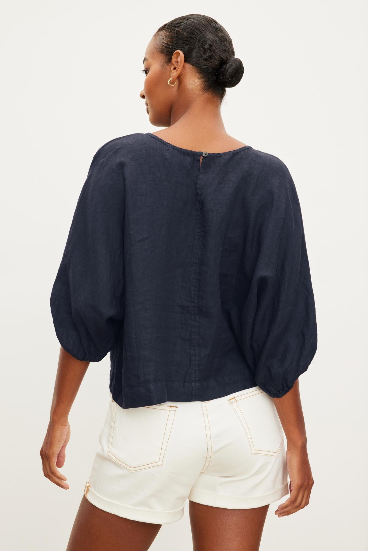   A person wearing a dark blue Velvet by Graham & Spencer CASS LINEN CREW NECK TOP and white shorts is shown from the back with their hair tied in a bun. 