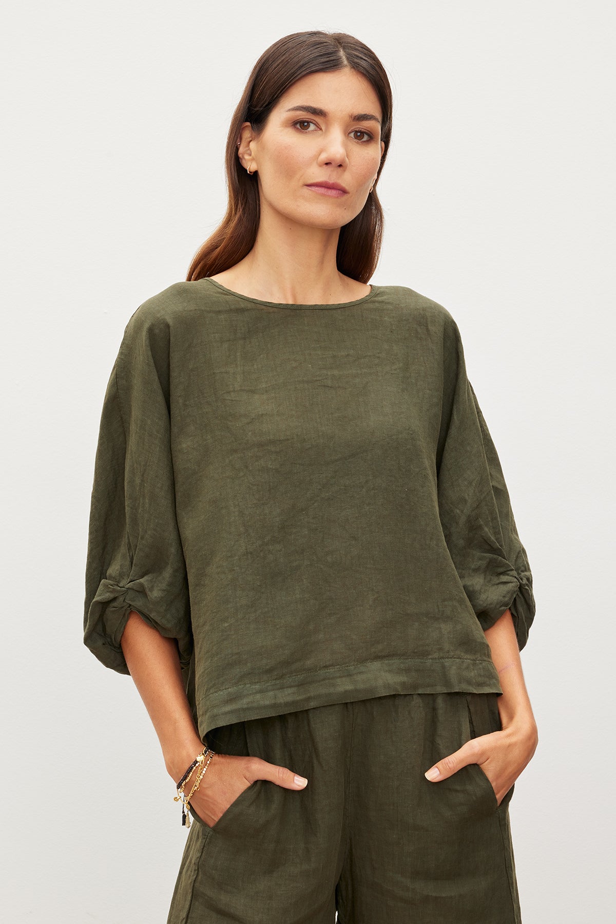 A person in the CASS LINEN TOP from Velvet by Graham & Spencer, featuring an olive green hue and a subtle twist knot detail, stands with hands in pockets against a plain background.-35955522830529