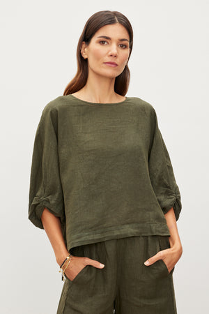 A person in the CASS LINEN TOP from Velvet by Graham & Spencer, featuring an olive green hue and a subtle twist knot detail, stands with hands in pockets against a plain background.