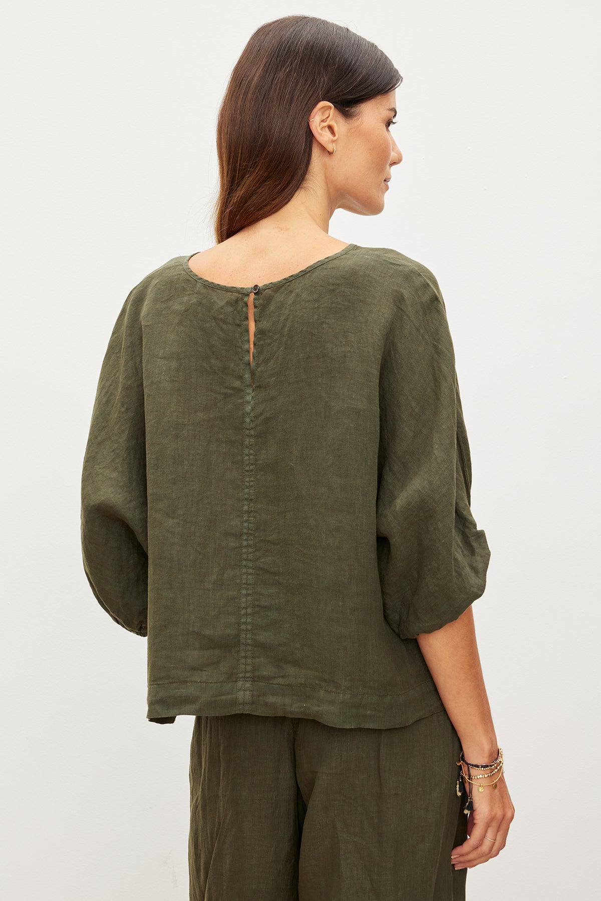 A woman stands with her back to the camera, wearing the CASS LINEN TOP and matching pants from Velvet by Graham & Spencer. The loose-fitting outfit's cocoon silhouette effortlessly enhances her style, offering a relaxed yet refined look in olive green.-35955522928833