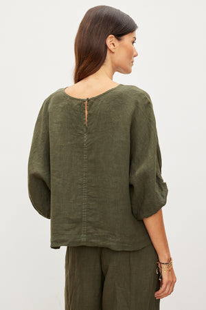 A woman stands with her back to the camera, wearing the CASS LINEN TOP and matching pants from Velvet by Graham & Spencer. The loose-fitting outfit's cocoon silhouette effortlessly enhances her style, offering a relaxed yet refined look in olive green.