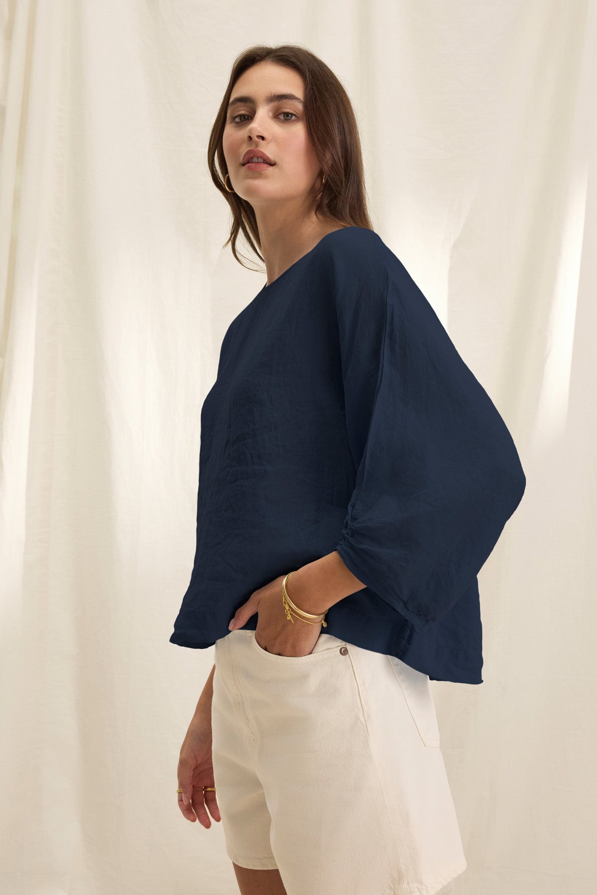 A woman stands before a cream-colored fabric backdrop, wearing the Velvet by Graham & Spencer CASS LINEN TOP—a loose, dark blue woven linen top with a round neck—paired with light beige shorts.