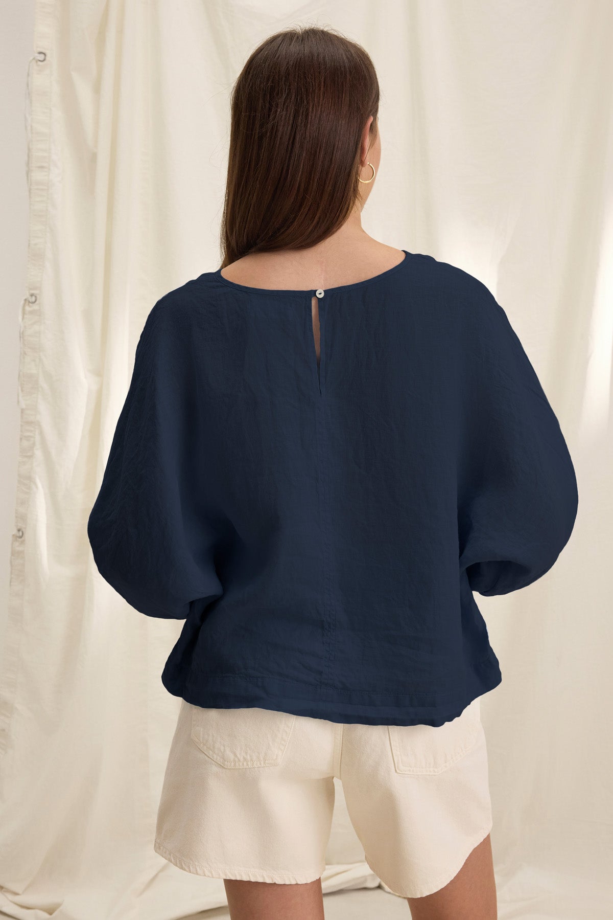 A woman faces away in front of a light fabric backdrop, wearing the CASS LINEN TOP by Velvet by Graham & Spencer in navy blue with 3/4 sleeves and cream shorts.-41160212054209