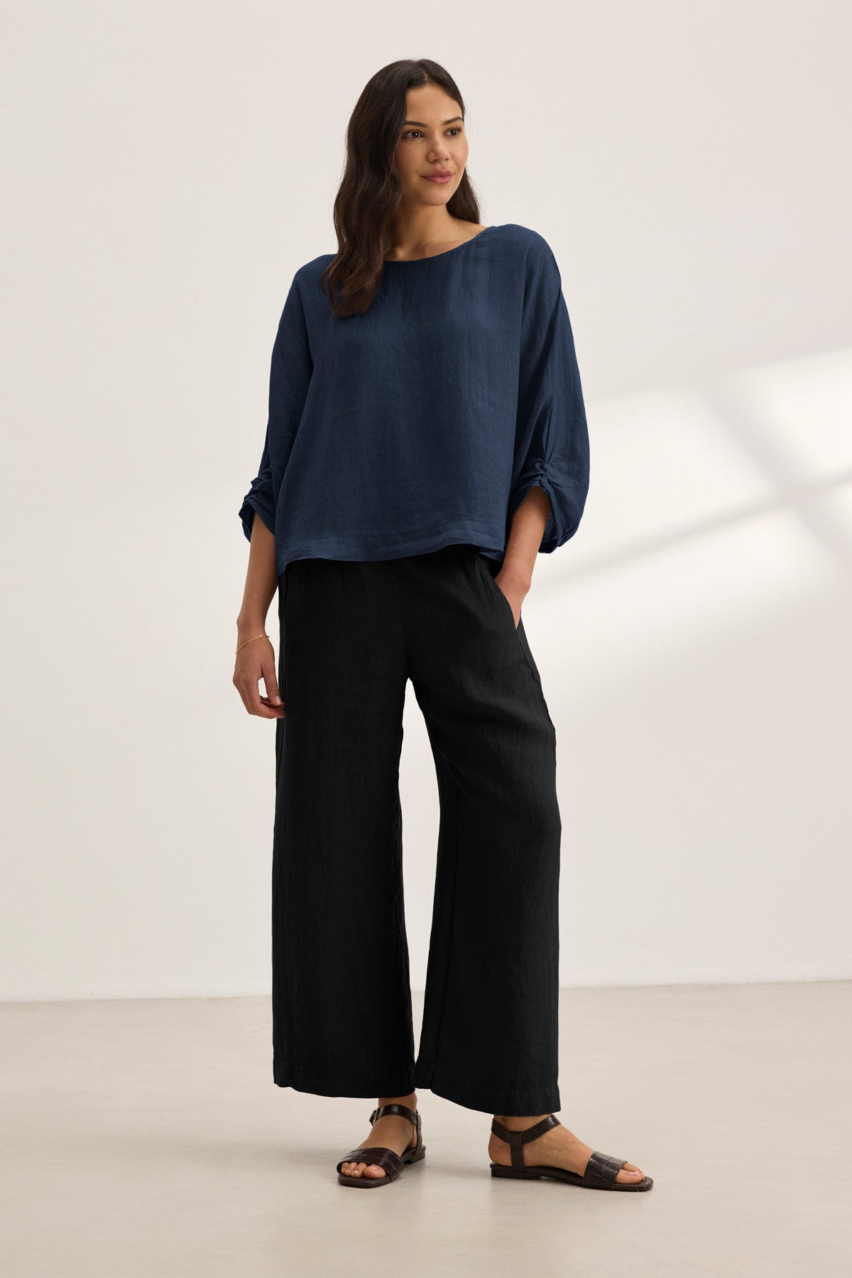 A woman stands in a minimal room wearing the CASS LINEN TOP by Velvet by Graham & Spencer, paired with black wide-leg pants and black sandals.-41263448883393