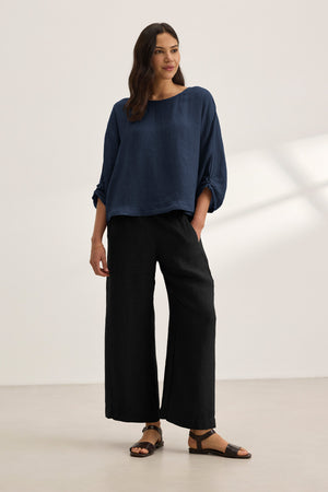 A woman stands in a minimal room wearing the CASS LINEN TOP by Velvet by Graham & Spencer, paired with black wide-leg pants and black sandals.
