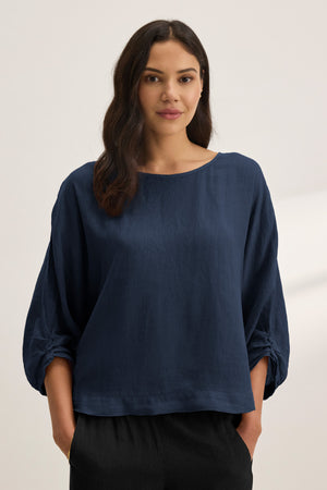 A woman stands facing forward in the CASS LINEN TOP by Velvet by Graham & Spencer—a loose-fitting navy blue woven linen top—paired with black pants against a plain light background.