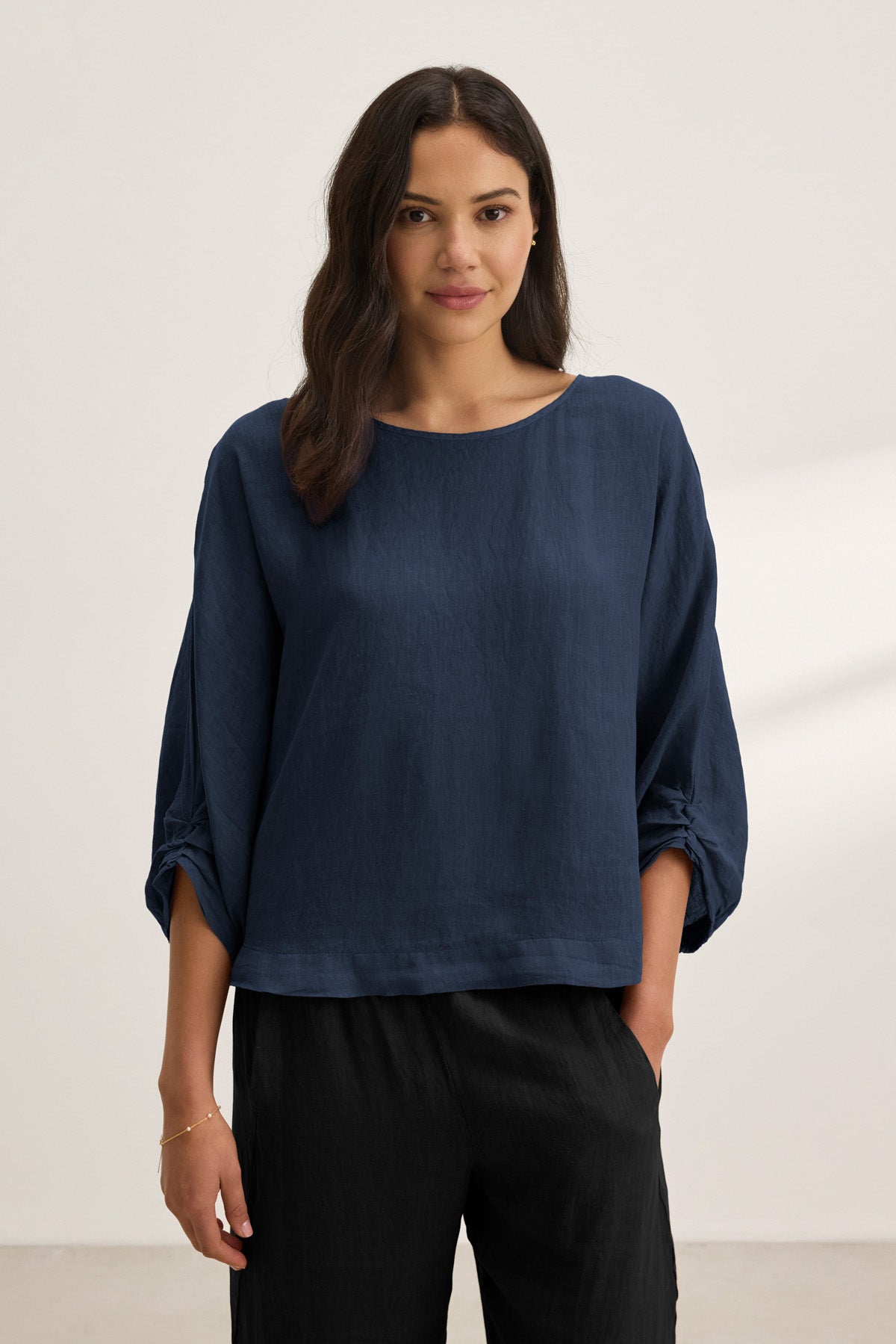 A woman with long dark hair wears the Velvet by Graham & Spencer CASS LINEN TOP in navy blue with 3/4 sleeves and black pants, standing against a plain light background.