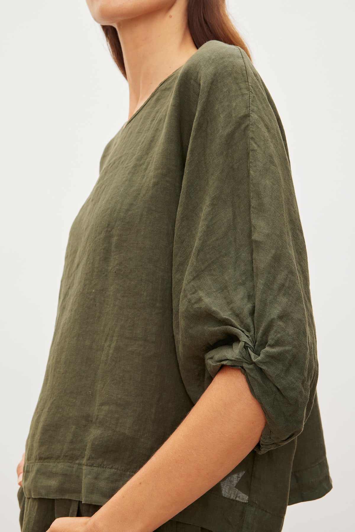   A person wearing the CASS LINEN TOP by Velvet by Graham & Spencer in olive green, featuring rolled-up sleeves and a subtle cocoon silhouette, facing to the right. 