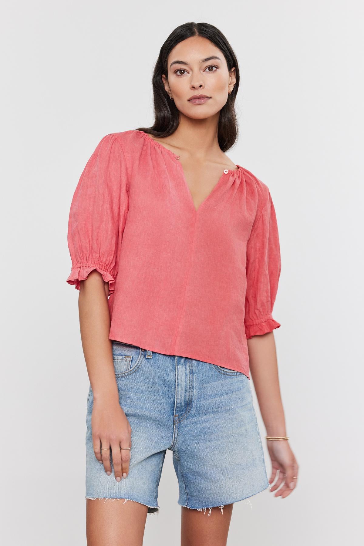   Woman wearing a Velvet by Graham & Spencer coral pink linen top with puff sleeves and denim shorts, standing against a white background. 