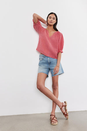 A woman in a pink Velvet by Graham & Spencer linen top with puff sleeves and denim shorts stands against a white backdrop, posing with one hand raised to her head.