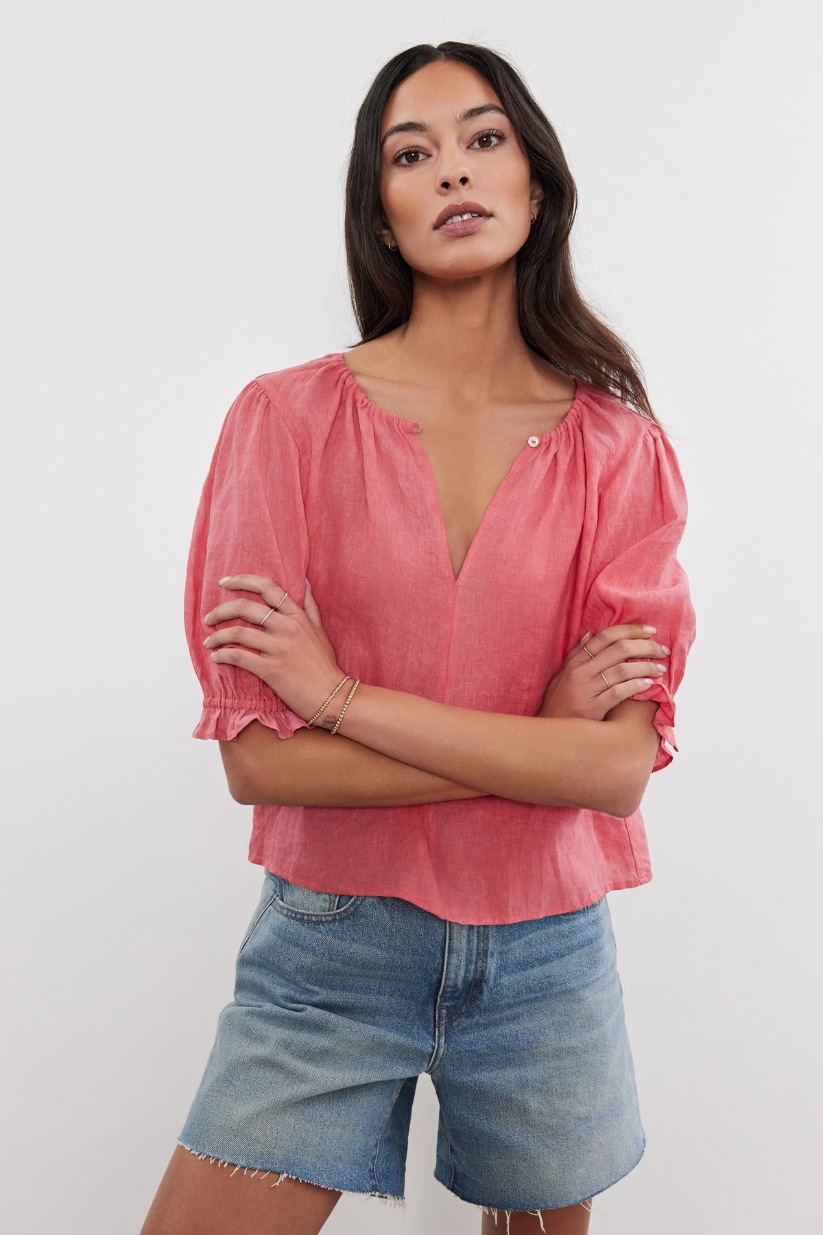   A woman stands confidently, wearing a Velvet by Graham & Spencer coral pink linen top with puff sleeves and denim shorts, with her arms crossed. She looks directly at the camera. 