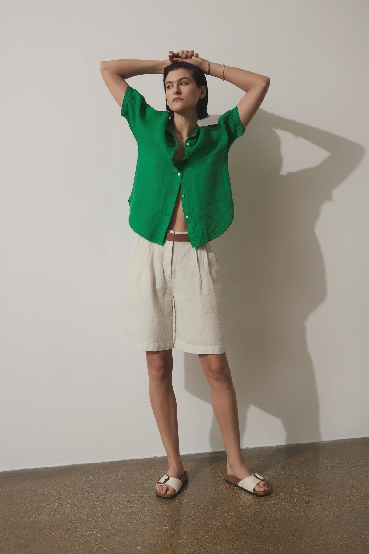   A woman stands against a plain wall, wearing a green button-up shirt, Velvet by Jenny Graham LARCHMONT HEAVY LINEN SHORTS, and white sandals, with her hands clasped above her head. 