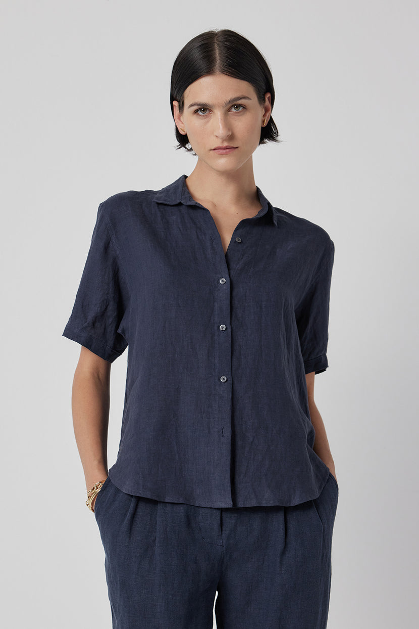 A woman in a CLAREMONT LINEN SHIRT by Velvet by Jenny Graham and matching pants stands against a light grey background, looking directly at the camera.