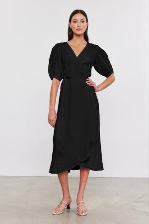 Woman standing in a studio, wearing a black Velvet by Graham & Spencer Dalene Linen Dress with puff sleeves and heeled sandals, posing for the camera.