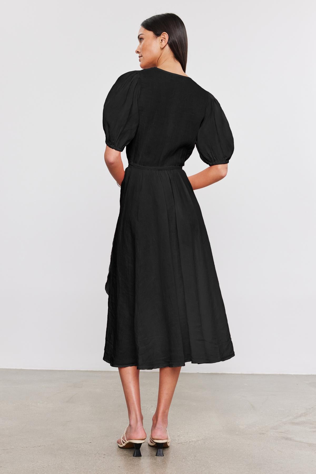   Woman in a black Velvet by Graham & Spencer Dalene Linen Dress with puffed sleeves standing, viewed from the back. She wears beige high heels and looks over her shoulder. 