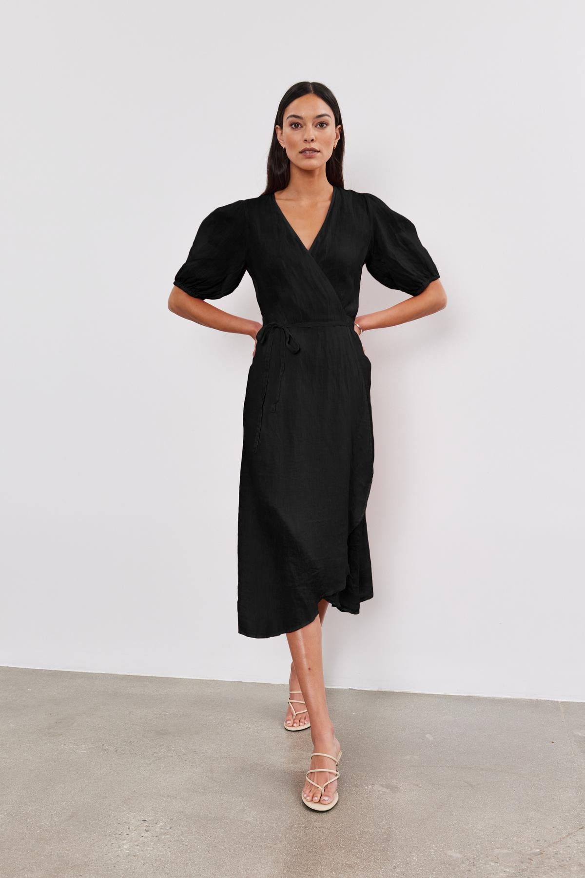 A woman stands against a white background, wearing a black v-neck wrap DALENE LINEN DRESS with short sleeves and a tie waist, paired with light-colored heeled sandals.-36917057978561