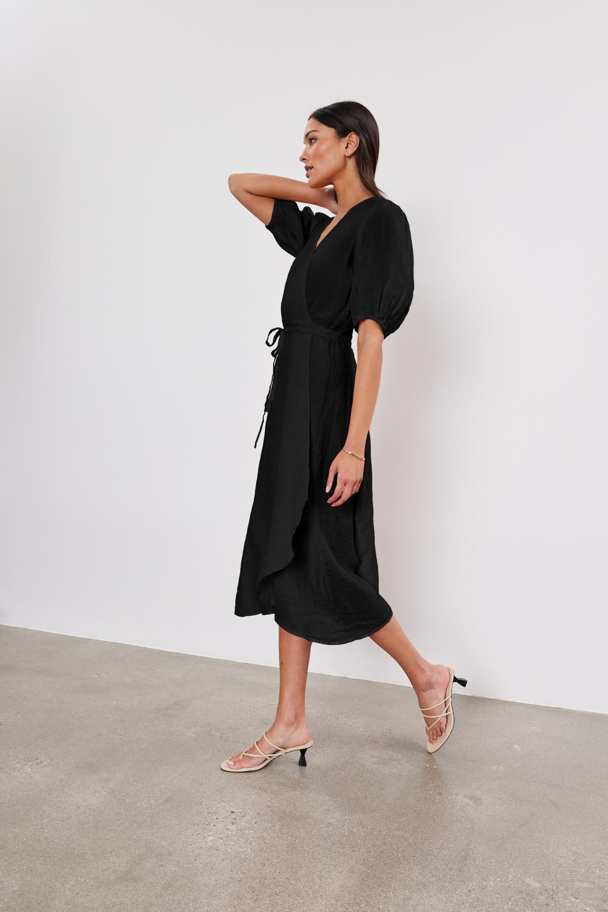   A woman in a black DaLENE LINEN DRESS with puffed sleeves walks in a minimalist space, looking to the side, wearing beige heels. Brand name: Velvet by Graham & Spencer 