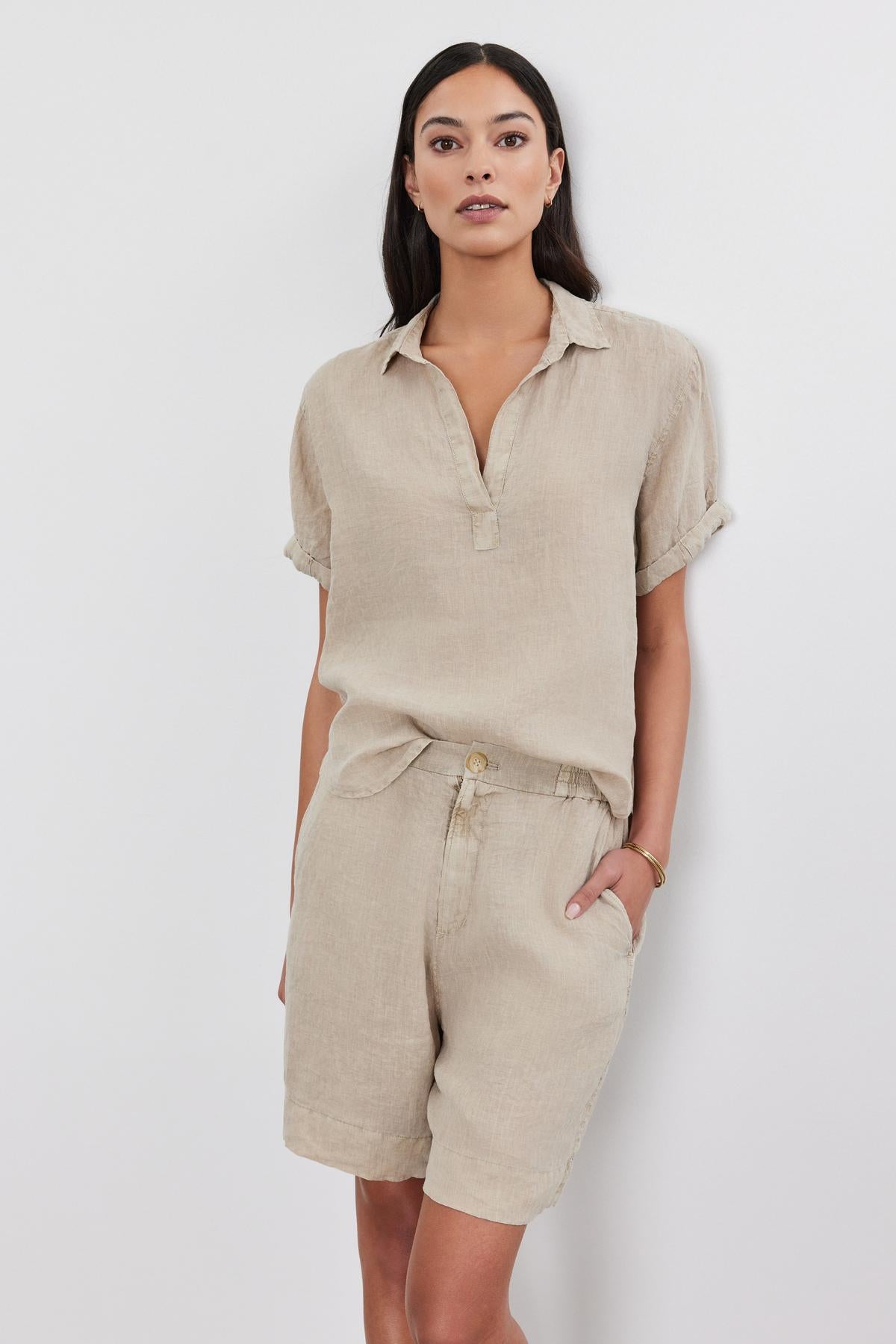   Woman wearing a beige short-sleeved breathable woven Velvet by Graham & Spencer linen shirt and matching shorts standing against a white background. 