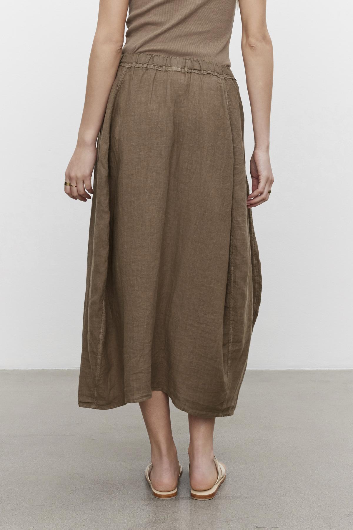 Woman standing in a brown mid-length FAE LINEN A-LINE SKIRT with an elastic waistband and beige shoes against a grey background by Velvet by Graham & Spencer.-36443407155393