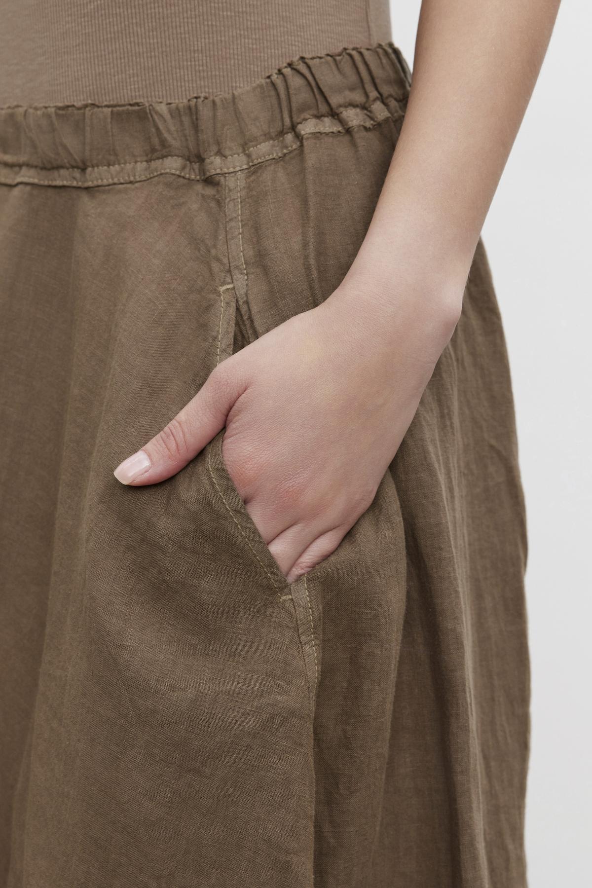   A close-up of a person's hand with fingers tucked into the pocket of a brown linen FAE LINEN A-LINE SKIRT by Velvet by Graham & Spencer. 