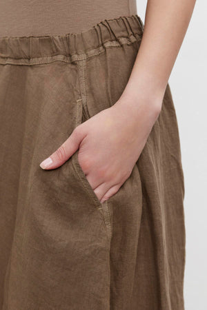 A close-up of a person's hand with fingers tucked into the pocket of a brown linen FAE LINEN A-LINE SKIRT by Velvet by Graham & Spencer.