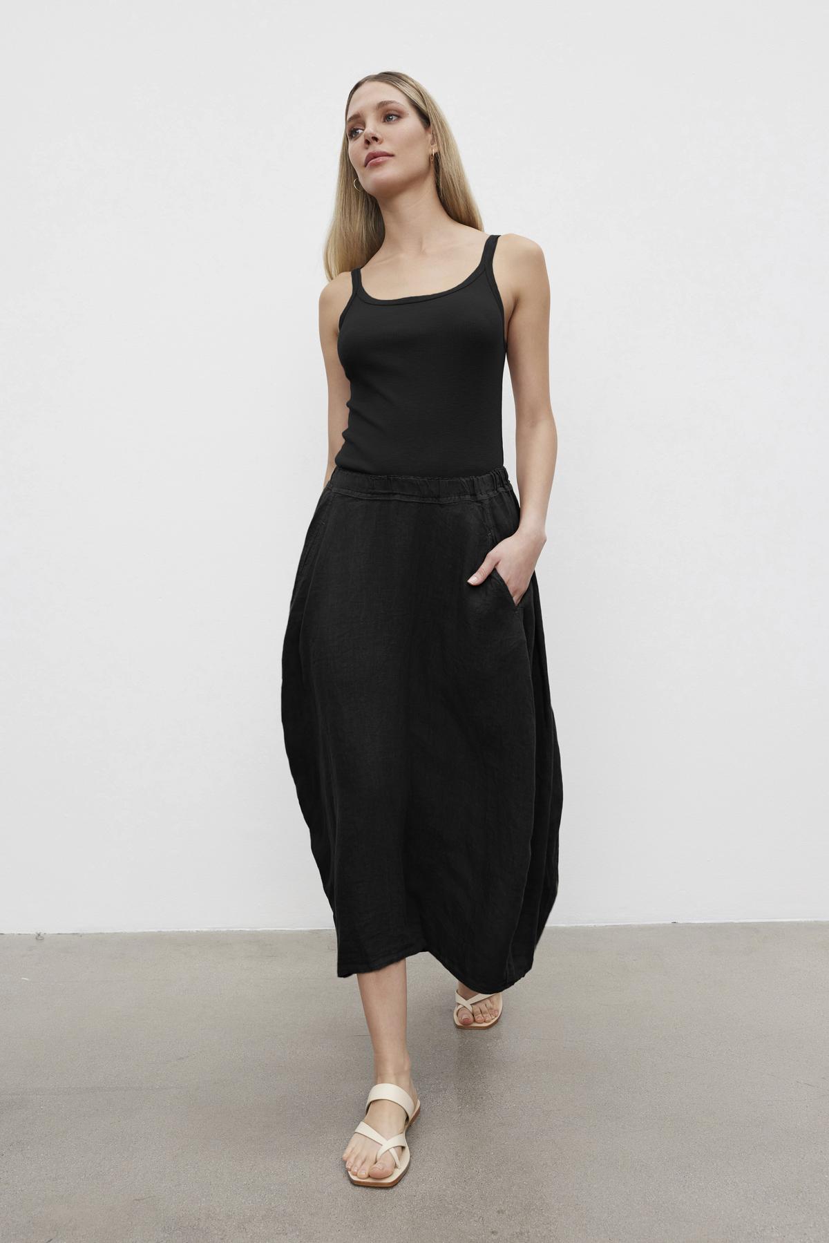   A person wearing a black ALIZA RIBBED COTTON LAYERING TANK by Velvet by Graham & Spencer, black high-waisted wide-leg pants, and beige sandals stands against a plain white background. They have long blonde hair and one hand in their pocket. 