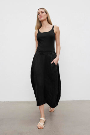 A person wearing a black ALIZA RIBBED COTTON LAYERING TANK by Velvet by Graham & Spencer, black high-waisted wide-leg pants, and beige sandals stands against a plain white background. They have long blonde hair and one hand in their pocket.