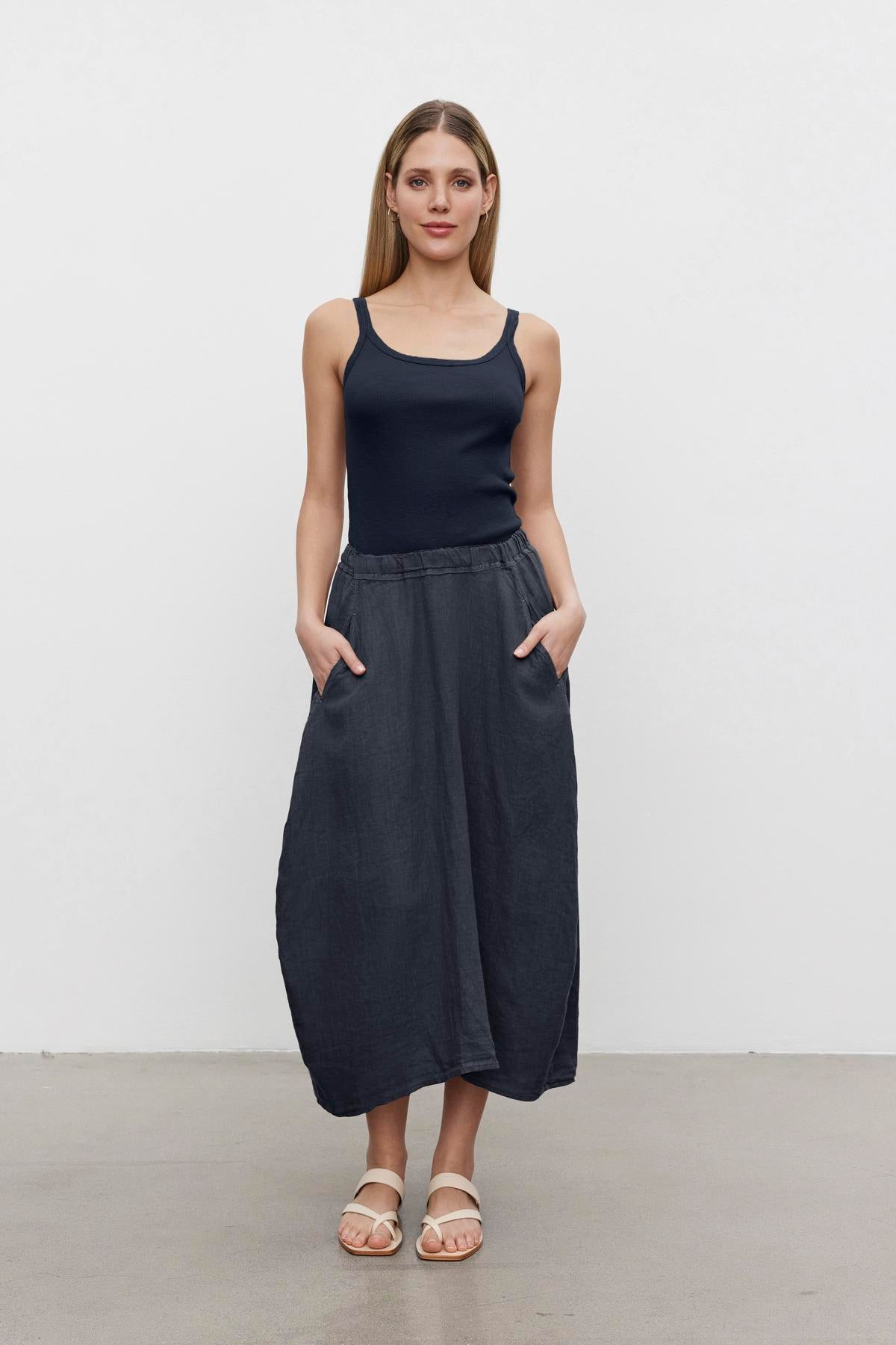   A woman stands against a plain white background, wearing a black ALIZA RIBBED COTTON LAYERING TANK by Velvet by Graham & Spencer with double-layer straps, a long dark skirt with pockets, and white sandals. 