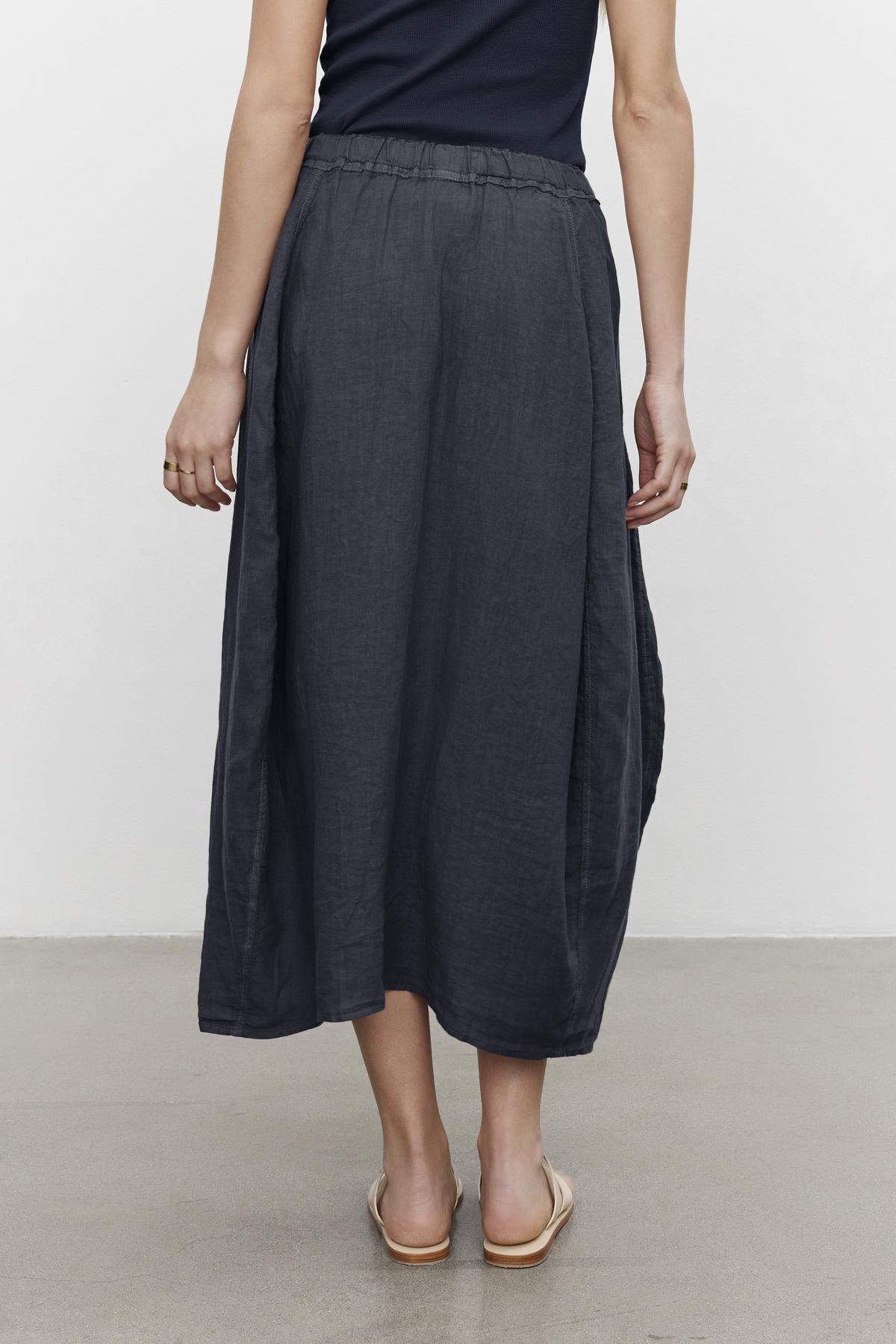   Woman viewed from behind wearing a dark gray FAE LINEN A-LINE SKIRT and light beige shoes, standing against a plain background by Velvet by Graham & Spencer. 