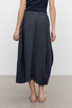 Woman viewed from behind wearing a dark gray FAE LINEN A-LINE SKIRT and light beige shoes, standing against a plain background by Velvet by Graham & Spencer.
