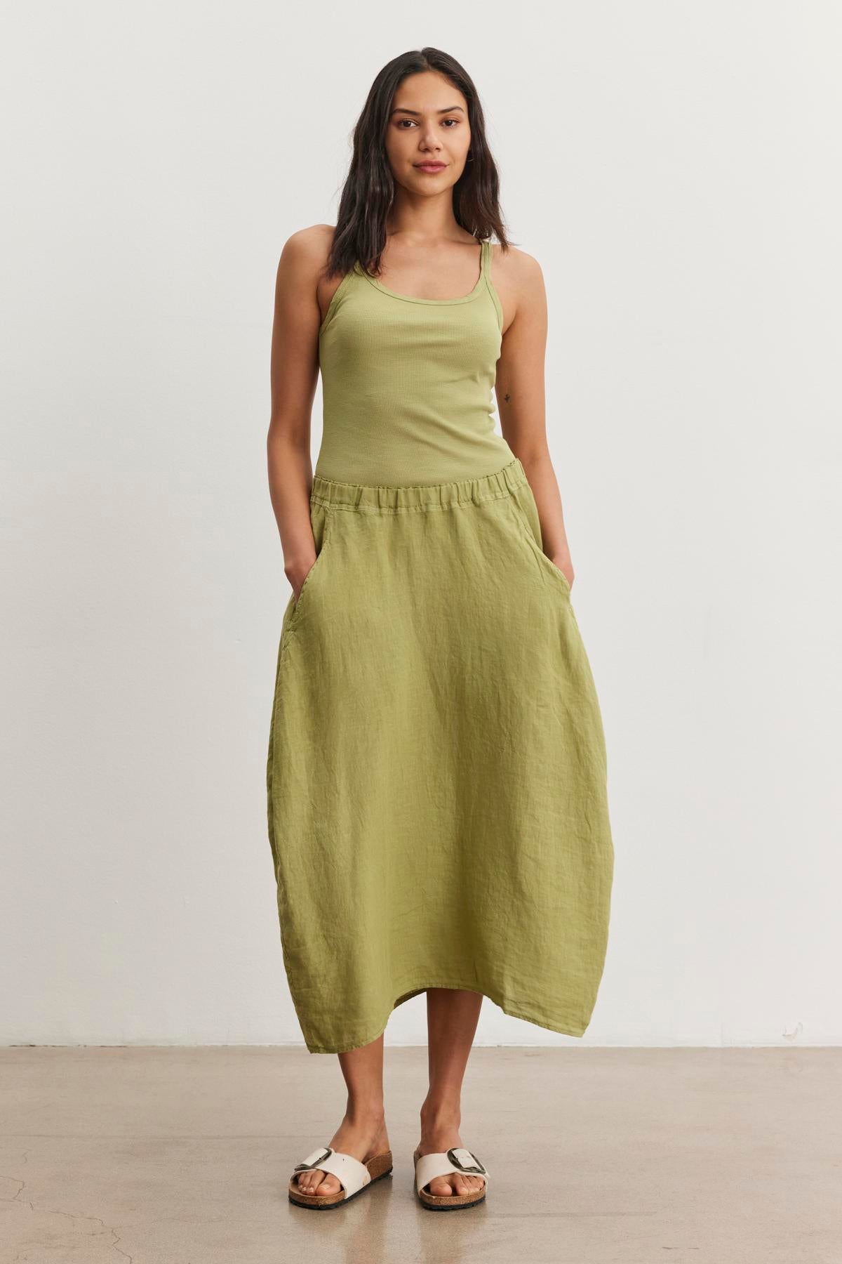   A woman stands against a plain background wearing a light green tank top and a matching A-line skirt with pockets, crafted from spring-weight linen, paired with white flat sandals. She is wearing the FAE LINEN SKIRT by Velvet by Graham & Spencer. 