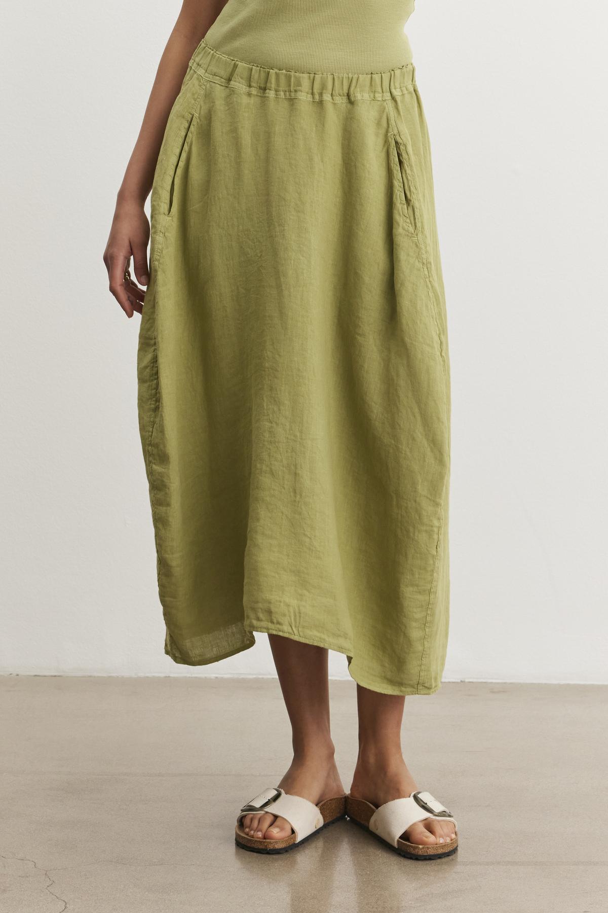   A person is wearing a light green, spring-weight FAE LINEN SKIRT by Velvet by Graham & Spencer with pockets and cream-colored sandals. The A-line skirt features an elastic waistband and offers a relaxed fit. They are standing on a light-colored floor. 