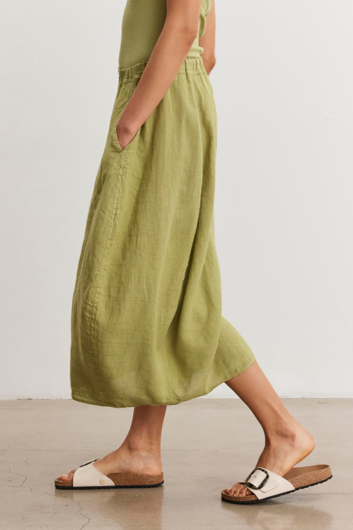   A person wearing a green FAE LINEN SKIRT by Velvet by Graham & Spencer with an elastic waistband and white sandals with cork soles walks on a light-colored floor. The person's hands are in the skirt pockets. 