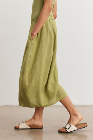 A person wearing a green FAE LINEN SKIRT by Velvet by Graham & Spencer with an elastic waistband and white sandals with cork soles walks on a light-colored floor. The person's hands are in the skirt pockets.