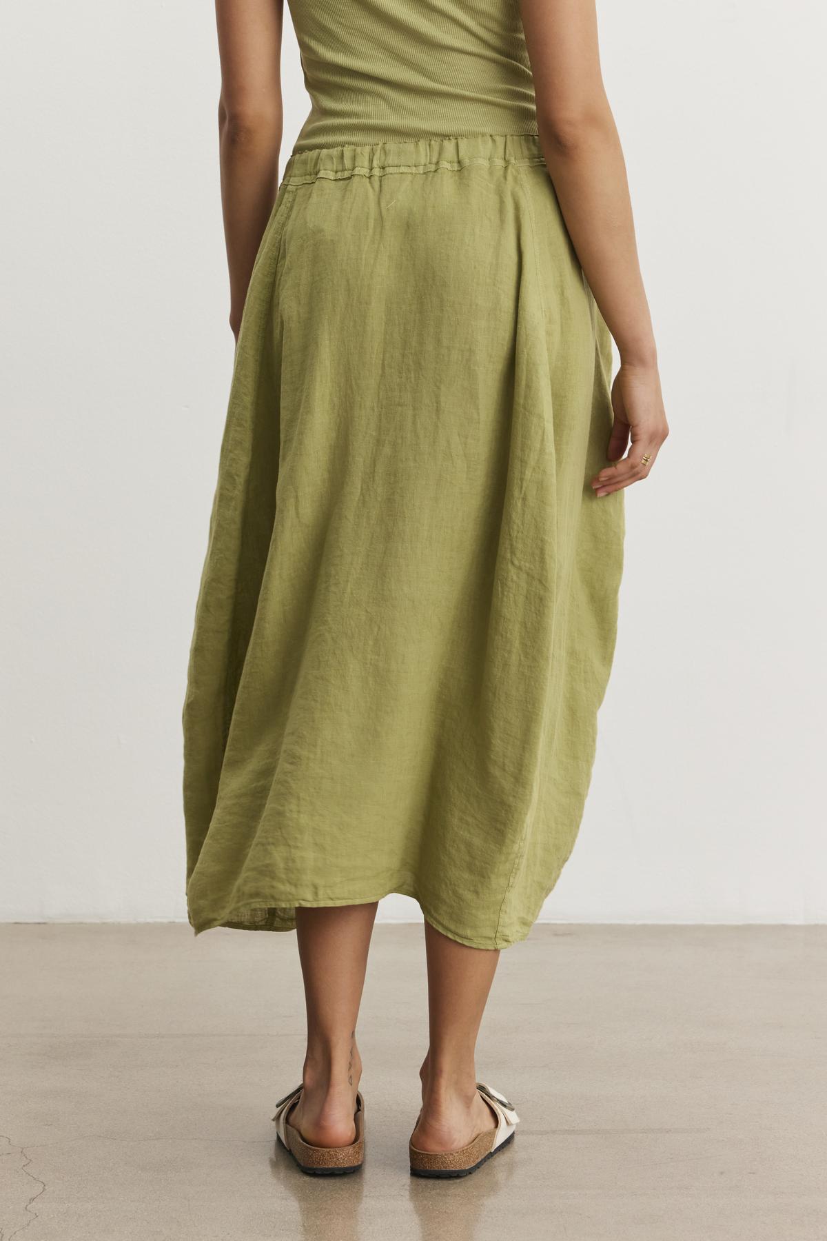   Woman wearing a green, spring-weight linen Velvet by Graham & Spencer FAE LINEN SKIRT with an elastic waistband and beige sandals, standing on a smooth, light-colored floor with a white background. Her upper body is partially visible. 