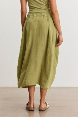 Woman wearing a green, spring-weight linen Velvet by Graham & Spencer FAE LINEN SKIRT with an elastic waistband and beige sandals, standing on a smooth, light-colored floor with a white background. Her upper body is partially visible.