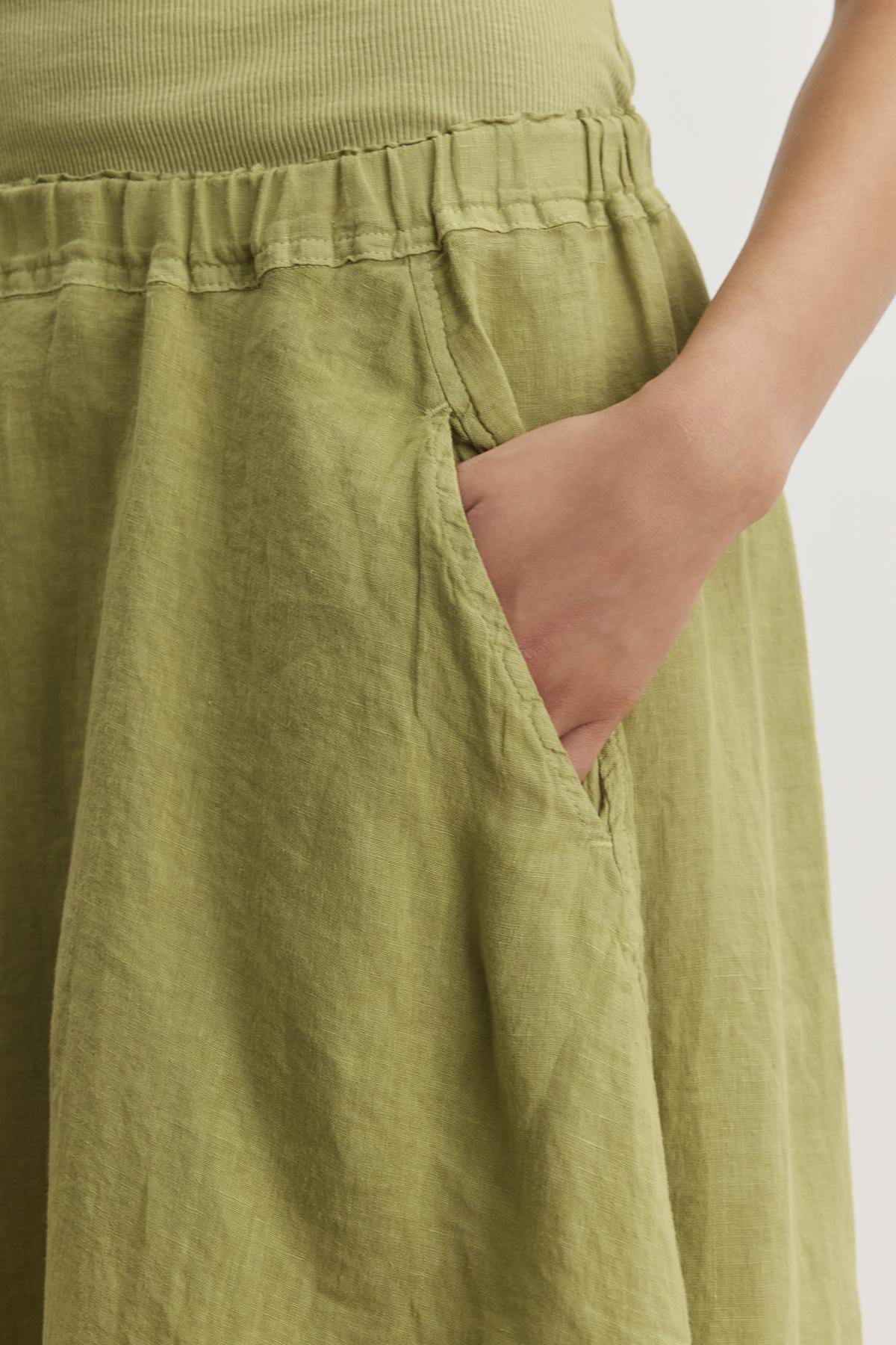   A person wearing a green FAE LINEN SKIRT with an elastic waistband, made from spring-weight linen, by Velvet by Graham & Spencer, with their right hand placed in one of the skirt's pockets. 