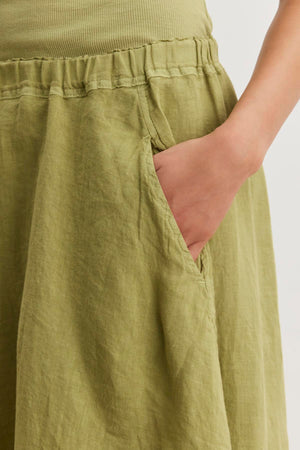 A person wearing a green FAE LINEN SKIRT with an elastic waistband, made from spring-weight linen, by Velvet by Graham & Spencer, with their right hand placed in one of the skirt's pockets.