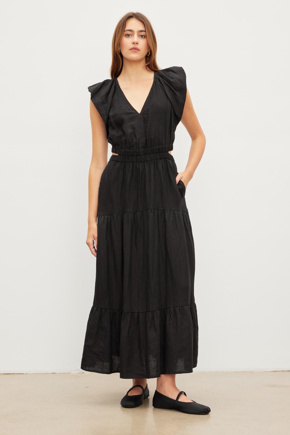 A woman stands against a plain background wearing the GINGER LINEN DRESS by Velvet by Graham & Spencer, which is a black breathable linen dress with a v-neckline and tiered skirt. She has her left hand in her pocket and is wearing black shoes.-35982607286465