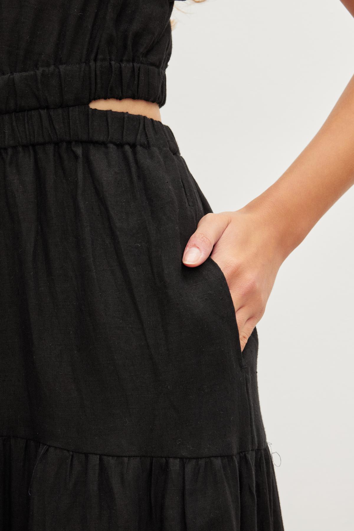   Close-up of a person wearing a black, breathable GINGER LINEN DRESS by Velvet by Graham & Spencer with an elastic waistband and v-neckline, and a hand resting in a side pocket. 