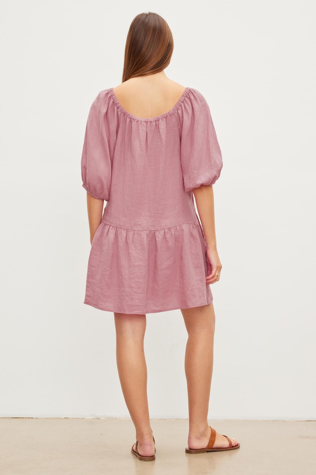   Woman viewed from behind wearing a pink, off-the-shoulder IRINA LINEN TIERED DRESS by Velvet by Graham & Spencer, with a ruffled hem and elastic neckline, standing against a plain background. 