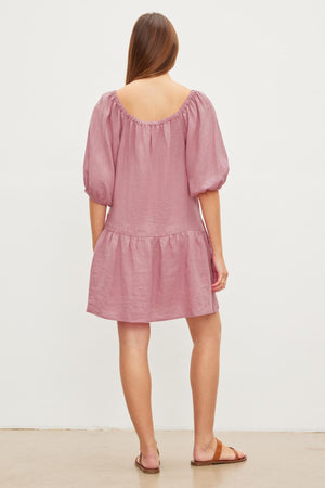 Woman viewed from behind wearing a pink, off-the-shoulder IRINA LINEN TIERED DRESS by Velvet by Graham & Spencer, with a ruffled hem and elastic neckline, standing against a plain background.
