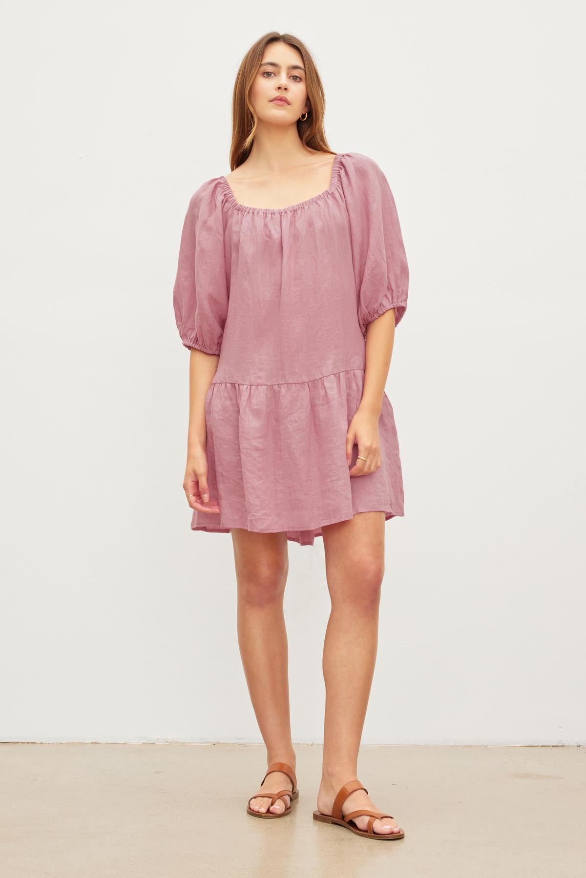   A woman stands against a plain background, wearing a pink off the shoulder IRINA LINEN TIERED DRESS with puff sleeves and brown sandals. Designed by Velvet by Graham & Spencer. 